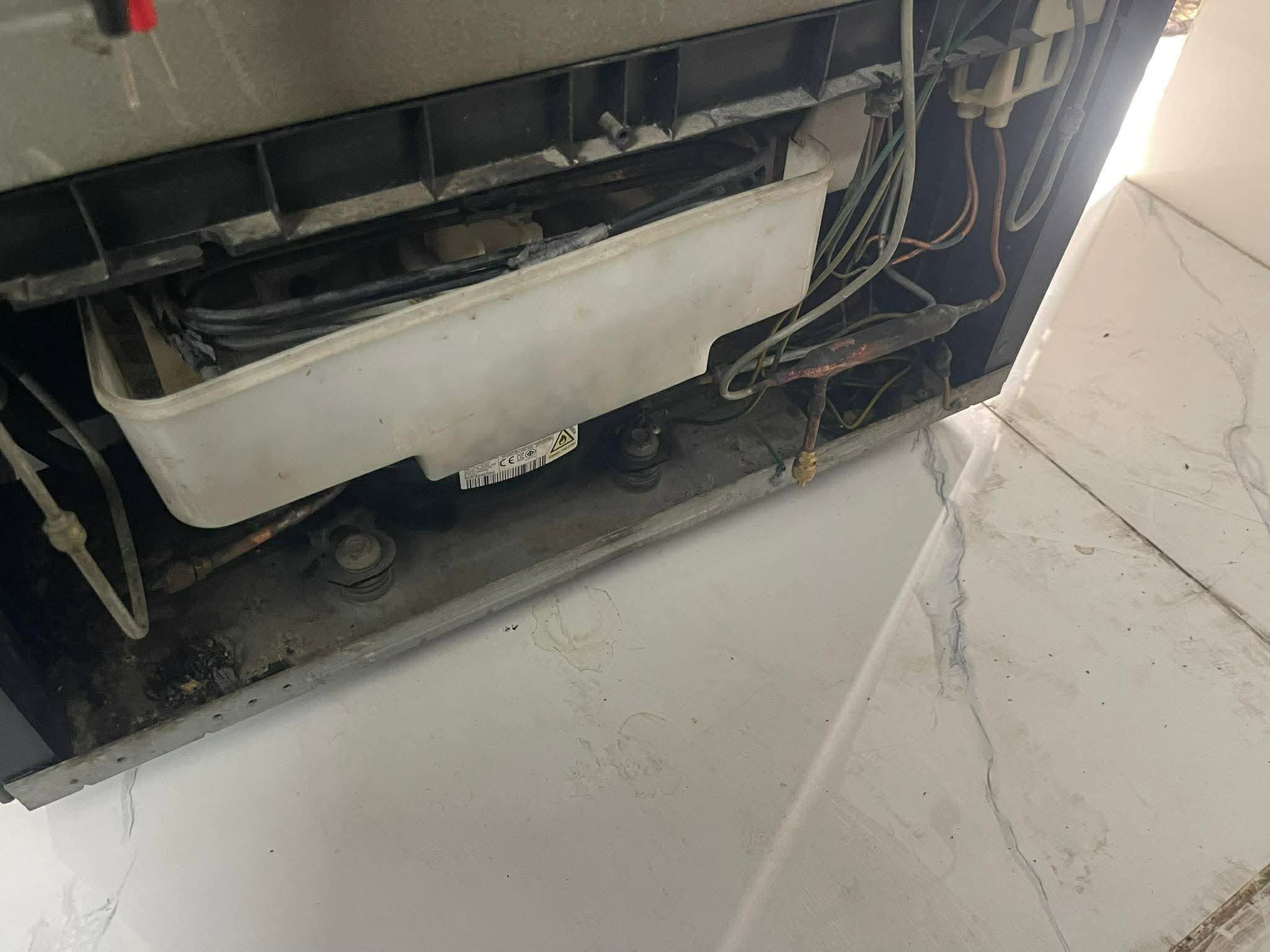 Gầm tủ lạnh mở nắp, lộ khay hứng nước xả đá, dây điện và đoạn ống đồng đã gỉ