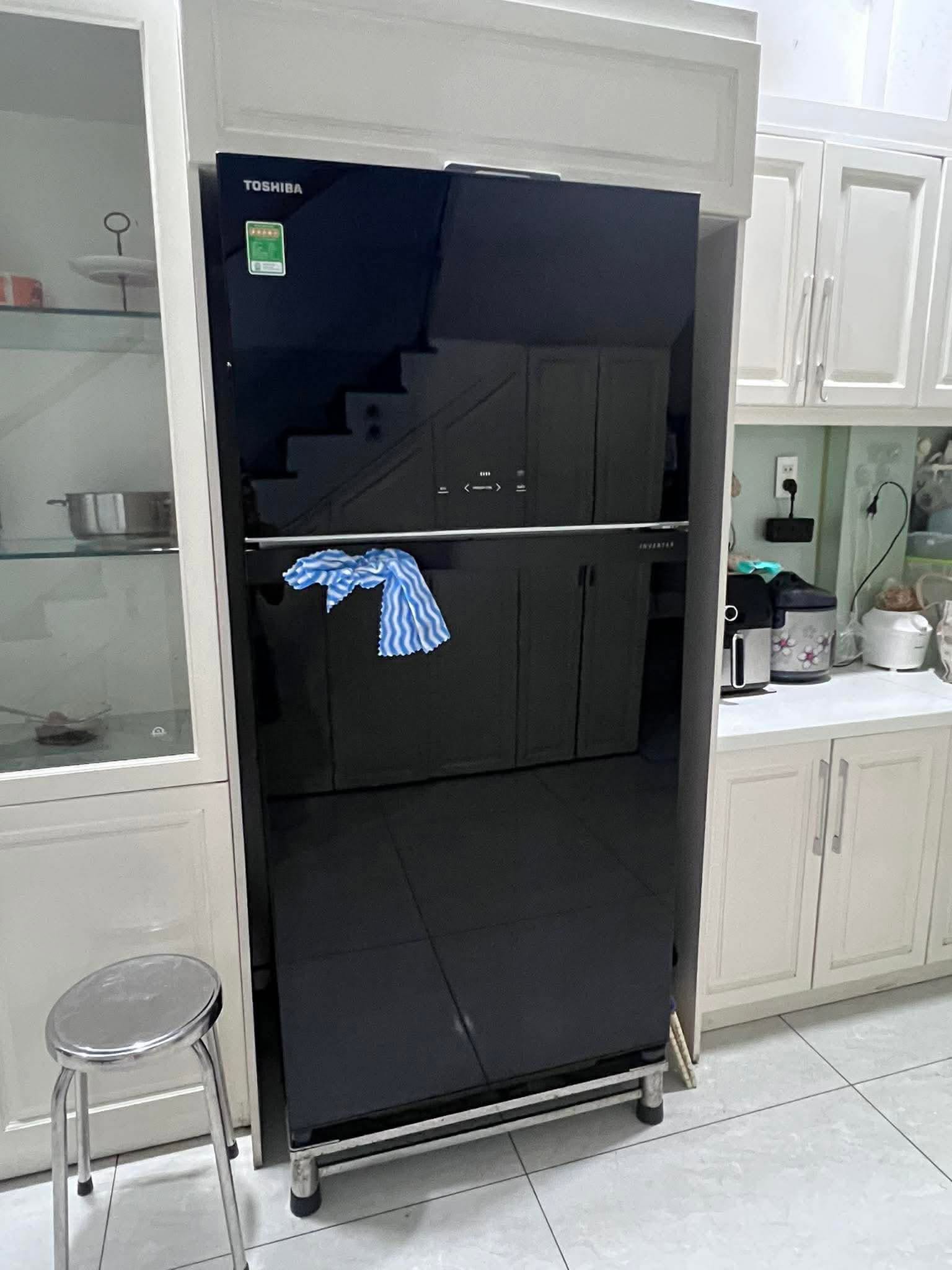 Tủ lạnh 2 cánh mặt kính đen đặt trên kệ chân, nằm gọn trong hốc tủ bếp màu trắng