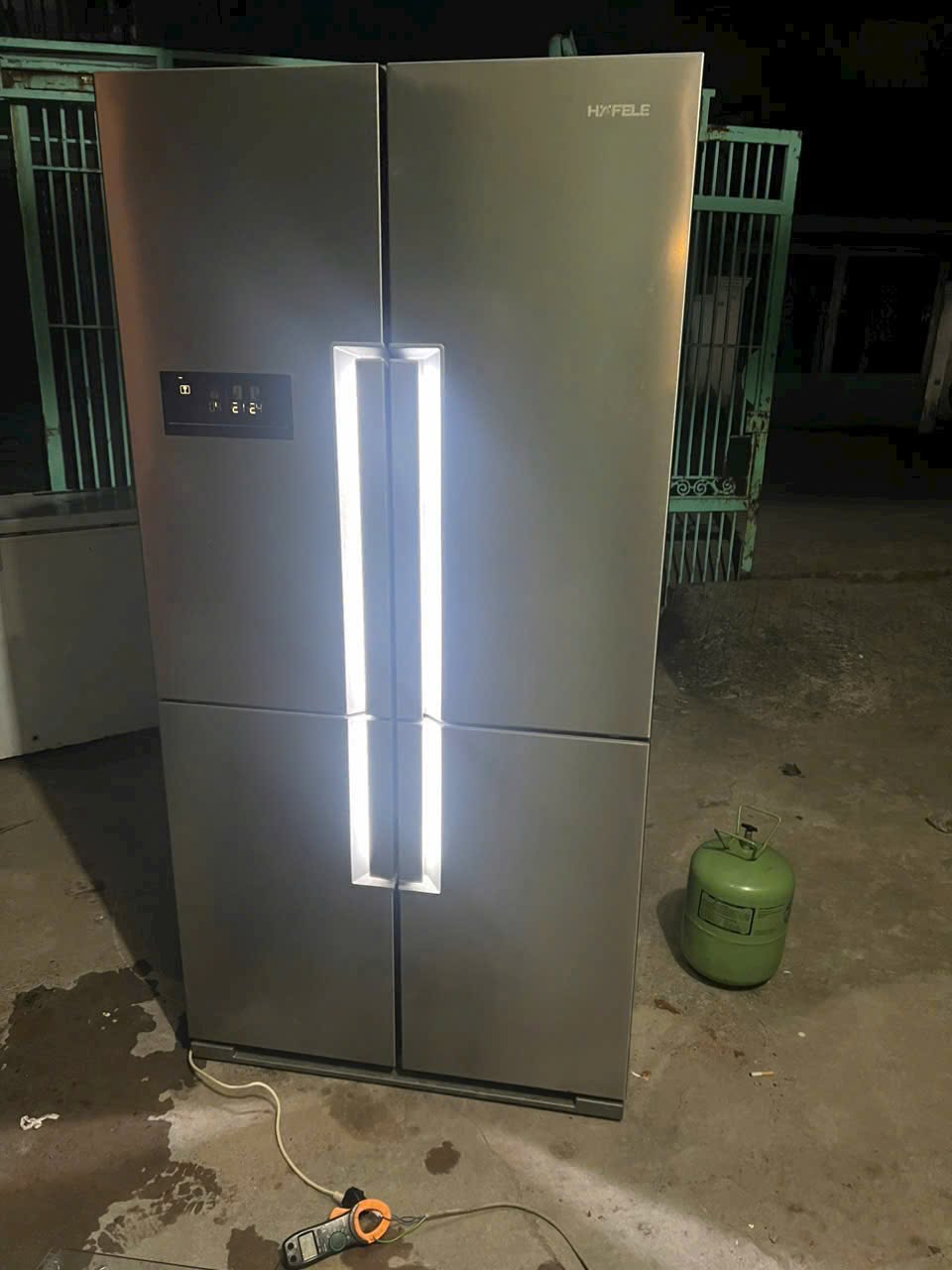 Tủ lạnh 4 cánh màu xám bật đèn chạy thử, bên cạnh có bình gas và ampe kìm