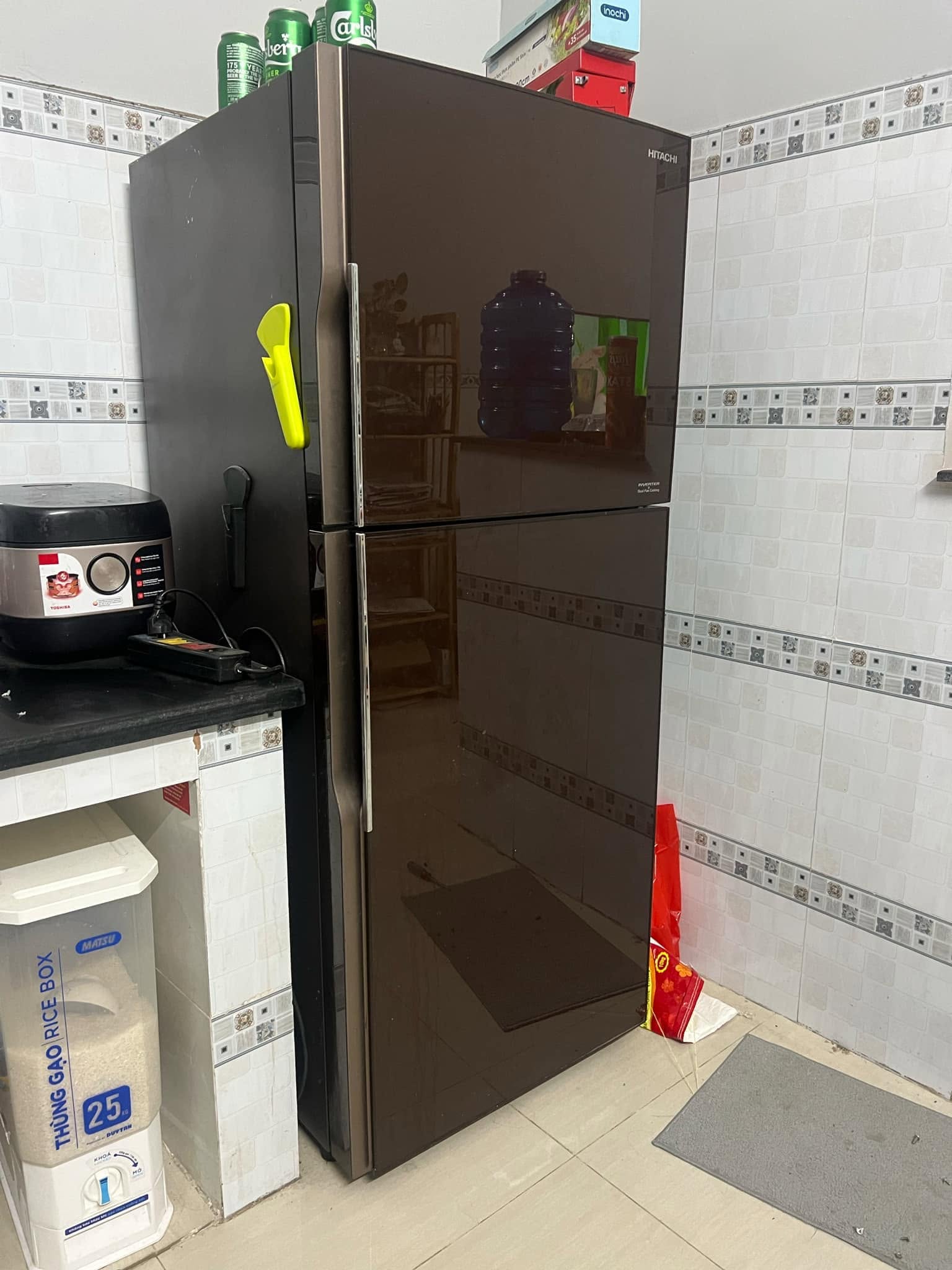 Tủ lạnh 2 cánh mặt gương nâu đặt trong góc bếp ốp gạch, nóc tủ chất thùng hàng và lon nước